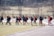 A group of individuals dressed in athletic clothing walk across a sports field. Many of them are carrying sports bags and some hold lacrosse sticks. They appear to be part of a team, wearing matching jackets and sneakers. The field is bordered by a fence and some trees in the background.