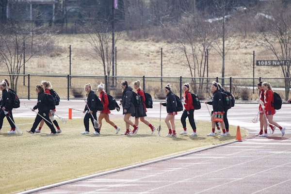 A group of individuals dressed in athletic clothing walk across a sports field. Many of them are carrying sports bags and some hold lacrosse sticks. They appear to be part of a team, wearing matching jackets and sneakers. The field is bordered by a fence and some trees in the background.