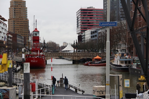 A bustling urban canal scene with a prominent red ship named 'VESSEL II' docked on the left side. Tall, modern buildings with distinctive architecture line both sides of the waterway, with a bridge visible in the background. The area is lively with pedestrians walking along the docks and several smaller boats visible on the water. A signpost with 'Leuvehaven' is in the foreground.