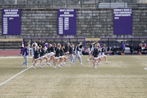 A group of athletes in sports attire are exercising or warming up on a grassy field. They are in a formation, appearing to be mid-stride, and the background shows a series of purple banners with sports achievements listed. The area is surrounded by a stone wall with benches and equipment visible.