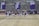 A group of athletes in sports attire are exercising or warming up on a grassy field. They are in a formation, appearing to be mid-stride, and the background shows a series of purple banners with sports achievements listed. The area is surrounded by a stone wall with benches and equipment visible.