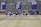 A group of athletes in sports attire are exercising or warming up on a grassy field. They are in a formation, appearing to be mid-stride, and the background shows a series of purple banners with sports achievements listed. The area is surrounded by a stone wall with benches and equipment visible.