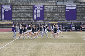 A group of athletes in sports attire are exercising or warming up on a grassy field. They are in a formation, appearing to be mid-stride, and the background shows a series of purple banners with sports achievements listed. The area is surrounded by a stone wall with benches and equipment visible.