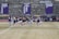 A group of athletes in sports attire are exercising or warming up on a grassy field. They are in a formation, appearing to be mid-stride, and the background shows a series of purple banners with sports achievements listed. The area is surrounded by a stone wall with benches and equipment visible.