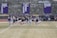A group of athletes in sports attire are exercising or warming up on a grassy field. They are in a formation, appearing to be mid-stride, and the background shows a series of purple banners with sports achievements listed. The area is surrounded by a stone wall with benches and equipment visible.