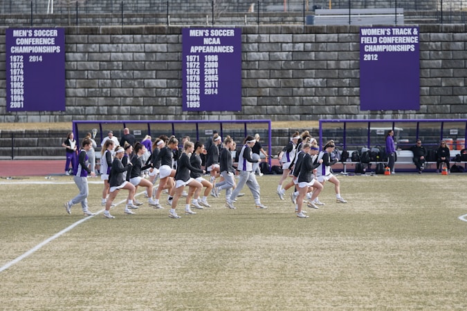 A group of athletes in sports attire are exercising or warming up on a grassy field. They are in a formation, appearing to be mid-stride, and the background shows a series of purple banners with sports achievements listed. The area is surrounded by a stone wall with benches and equipment visible.