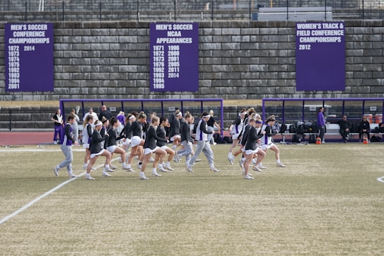 A group of athletes in sports attire are exercising or warming up on a grassy field. They are in a formation, appearing to be mid-stride, and the background shows a series of purple banners with sports achievements listed. The area is surrounded by a stone wall with benches and equipment visible.