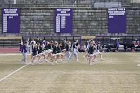 A group of athletes in sports attire are exercising or warming up on a grassy field. They are in a formation, appearing to be mid-stride, and the background shows a series of purple banners with sports achievements listed. The area is surrounded by a stone wall with benches and equipment visible.