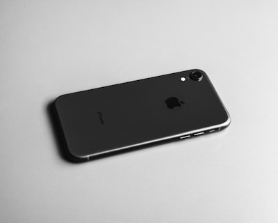 A black smartphone is displayed on its back, showcasing its sleek and minimalist design. The camera lens and a small flash are located at the top corner of the device. The surface has a smooth matte finish, and an iconic logo is centered on the phone.