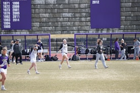 A group of athletes are on a sports field with a purple and white color scheme. Some appear to be warming up or stretching, while others are gathered near a bench. A large scoreboard or banner displaying years is visible in the background.