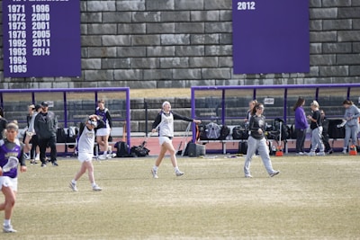 A group of athletes are on a sports field with a purple and white color scheme. Some appear to be warming up or stretching, while others are gathered near a bench. A large scoreboard or banner displaying years is visible in the background.