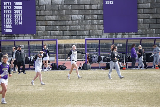 A group of athletes are on a sports field with a purple and white color scheme. Some appear to be warming up or stretching, while others are gathered near a bench. A large scoreboard or banner displaying years is visible in the background.