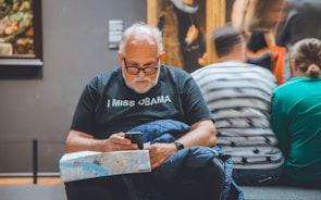 An older man with white hair and a beard looks intently at his smartphone. He wears glasses and a black t-shirt with the text 'I MISS ØBAMA'. In his lap is a folded map and a blue jacket. He's seated in a gallery or museum setting, with blurred figures and paintings visible in the background.
