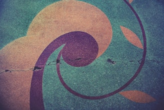 A cracked concrete surface features colorful, swirling geometric patterns incorporating circles, arcs, and leaf-like shapes. The design contrasts against the aged, slightly rough texture of the pavement.