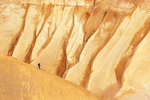 A lone person is hiking across a vast, barren landscape composed of steep, eroded formations with layered shades of brown and tan. The terrain appears to be a natural earth formation, possibly a canyon or desert badlands.