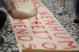 A person is painting red letters on a large piece of paper placed on a stone walkway. The letters appear to be part of a message or protest sign. Beside the painter, several people stand around wearing casual clothing, suggesting a public event or gathering.