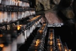 Rows of lit votive candles are arranged on metal racks with a stone wall backdrop, creating a serene and contemplative ambiance.