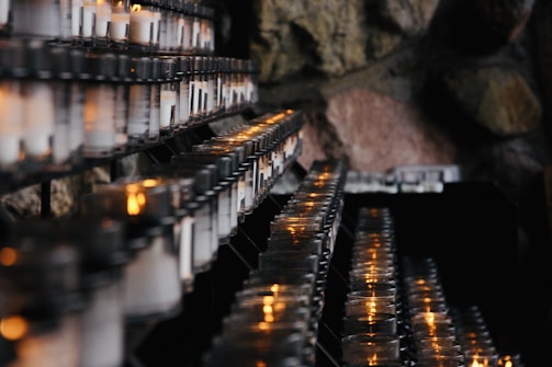 Rows of lit votive candles are arranged on metal racks with a stone wall backdrop, creating a serene and contemplative ambiance.