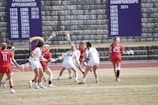 A group of female lacrosse players in action on a field. Players are wearing red and white uniforms and are engaged in the game, holding sticks and focusing on the ball. There is a stone wall in the background with banners displaying championship and postseason appearances.