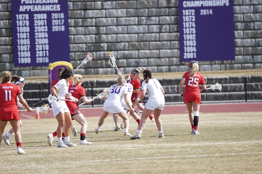 A group of female lacrosse players in action on a field. Players are wearing red and white uniforms and are engaged in the game, holding sticks and focusing on the ball. There is a stone wall in the background with banners displaying championship and postseason appearances.