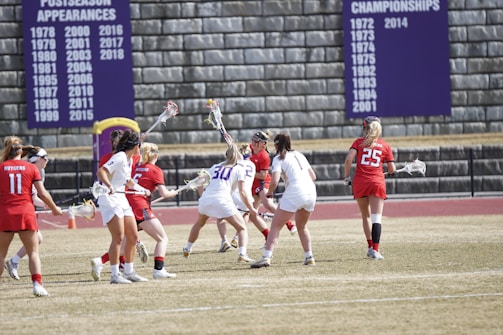 A group of female lacrosse players in action on a field. Players are wearing red and white uniforms and are engaged in the game, holding sticks and focusing on the ball. There is a stone wall in the background with banners displaying championship and postseason appearances.