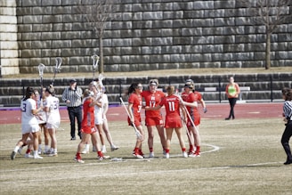 A group of female lacrosse players are gathered on a sports field. They are wearing uniforms in red and white, representing opposing teams. Some players hold lacrosse sticks high, indicating a moment of celebration or discussion. Referees in striped shirts are present, and a stone wall forms the background with a track running behind the field.