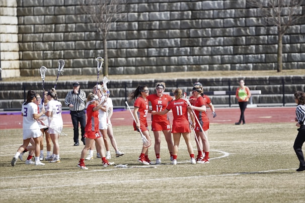 A group of female lacrosse players are gathered on a sports field. They are wearing uniforms in red and white, representing opposing teams. Some players hold lacrosse sticks high, indicating a moment of celebration or discussion. Referees in striped shirts are present, and a stone wall forms the background with a track running behind the field.