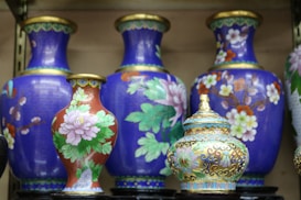 A collection of intricately designed ceramic vases and a lidded pot are displayed together. The vases mostly feature a vibrant blue background adorned with colorful floral patterns, including blossoms in white, pink, and orange hues. The lidded pot is smaller, showcasing ornate details with gold accents and floral designs.