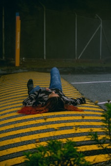 A person is lying on a road with yellow and black striped speed bumps. The individual is wearing jeans and a jacket, with long hair splayed out. Surrounding the scene are dark, overcast conditions with trees visible in the background.