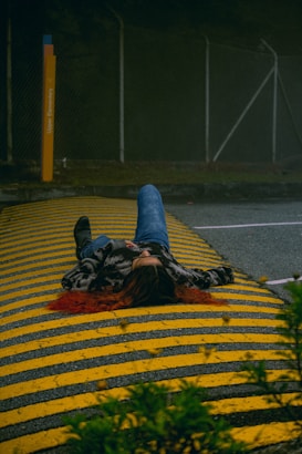 A person is lying on a road with yellow and black striped speed bumps. The individual is wearing jeans and a jacket, with long hair splayed out. Surrounding the scene are dark, overcast conditions with trees visible in the background.