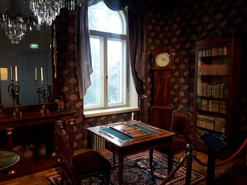 An elegant, dimly-lit room with vintage furniture, including a wooden table set for a game, surrounded by chairs. The upholstery and wallpaper have rich patterns, and a large window lets in soft light. On the wall is a tall clock, and a bookshelf filled with old books. A chandelier hangs from the ceiling, adding a luxurious touch.