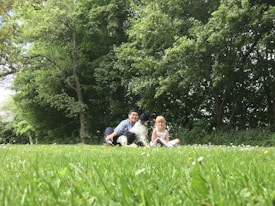 A peaceful outdoor scene with a man, a child, and a black and white dog sitting on a grassy field. The background is filled with lush green trees and small wildflowers scattered across the grass.