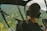 A person wearing headphones is seated inside a helicopter, looking out at a lush, green landscape from the cockpit. The view includes mountain ridges and valleys, with some small buildings visible below. Various controls and instruments are visible inside the cockpit.