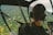 A person wearing headphones is seated inside a helicopter, looking out at a lush, green landscape from the cockpit. The view includes mountain ridges and valleys, with some small buildings visible below. Various controls and instruments are visible inside the cockpit.