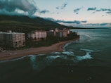 A tranquil seaside view with several large resort buildings lining a sandy beach as gentle waves roll onto the shore. The scene is set against a backdrop of lush, green mountains partially shrouded by clouds. The sky is a mix of blues and whites, indicating early morning or late afternoon light.