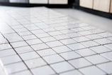 A close-up view of a grid pattern formed by a series of small, shiny white ceramic tiles. The surface appears clean and polished, with light reflecting softly off the tiles, creating a slightly glossy effect. The background is blurred, which emphasizes the uniform arrangement and texture of the tiles.