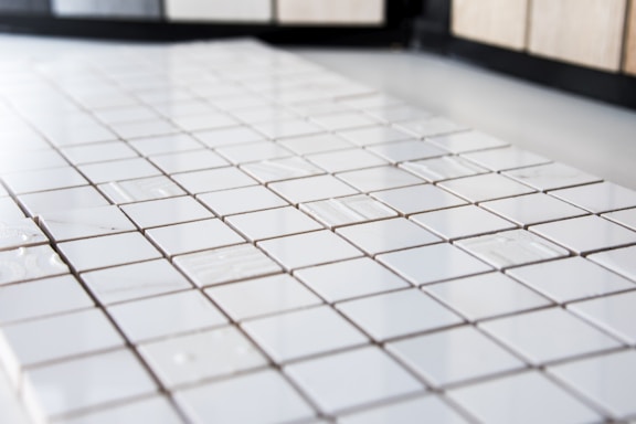 A close-up view of a grid pattern formed by a series of small, shiny white ceramic tiles. The surface appears clean and polished, with light reflecting softly off the tiles, creating a slightly glossy effect. The background is blurred, which emphasizes the uniform arrangement and texture of the tiles.