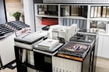 A room filled with organized catalogs and books, many stacked on tables and shelves. The shelves are lined with various binders and samples. A small green potted plant is visible in the background.