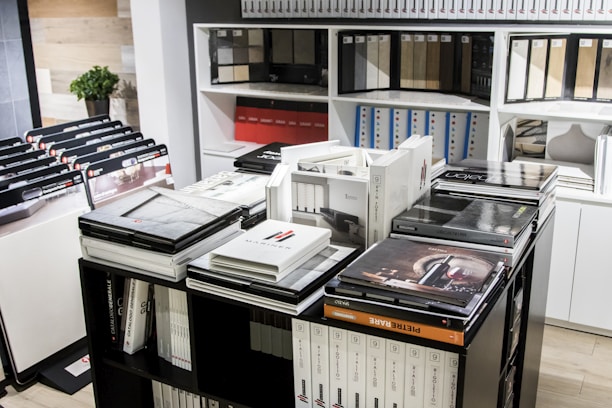 A room filled with organized catalogs and books, many stacked on tables and shelves. The shelves are lined with various binders and samples. A small green potted plant is visible in the background.