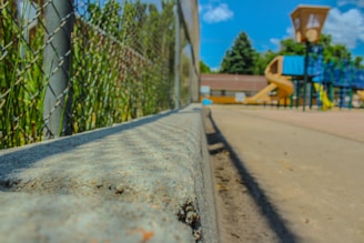 A playground with a focus on a chain-link fence and concrete barrier leading towards colorful playground equipment. A slide and play structures are set against a background of trees under a bright blue sky with occasional clouds.
