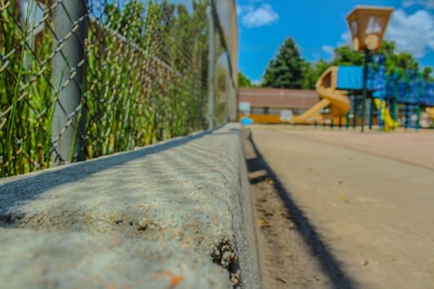 A playground with a focus on a chain-link fence and concrete barrier leading towards colorful playground equipment. A slide and play structures are set against a background of trees under a bright blue sky with occasional clouds.