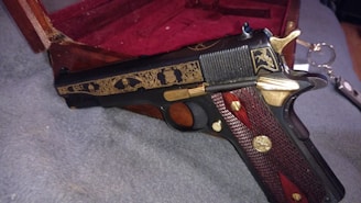 A decorative handgun with intricate gold engravings rests on a textured grey surface. The wood and metal handle features detailed patterns, and there is a wooden box with a red velvet lining partially visible in the background. A set of keys lies nearby.