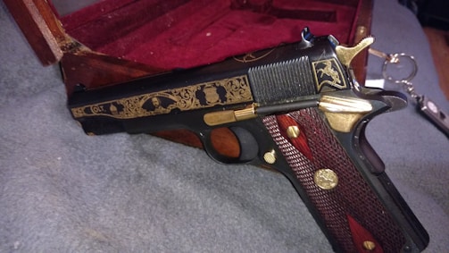 A decorative handgun with intricate gold engravings rests on a textured grey surface. The wood and metal handle features detailed patterns, and there is a wooden box with a red velvet lining partially visible in the background. A set of keys lies nearby.