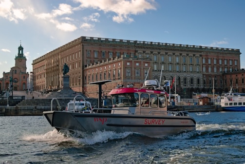 A survey boat navigates through choppy waters against the backdrop of an imposing historical building with numerous windows. A statue stands prominently in the foreground, and a church with a tall spire is visible to the left. The sky is partly cloudy, casting a serene light over the scene.