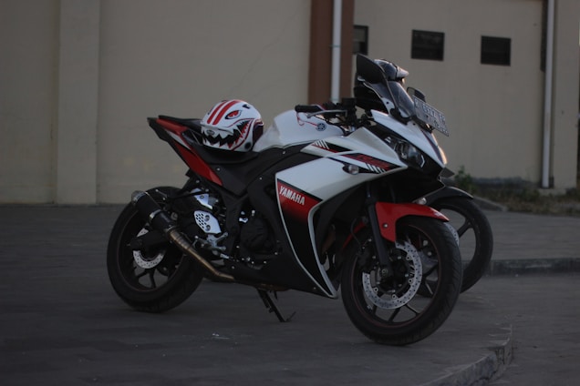 A sports motorcycle is parked on a paved surface with a textured background. The bike features a sleek design with prominent branding and a combination of black, red, and white colors. On top of the motorcycle seat, a helmet with a stylized design is placed.