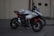 A sports motorcycle is parked on a paved surface with a textured background. The bike features a sleek design with prominent branding and a combination of black, red, and white colors. On top of the motorcycle seat, a helmet with a stylized design is placed.