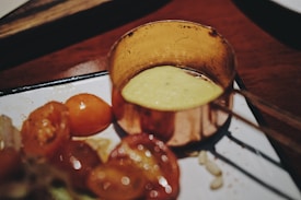 A small metal pot filled with a yellow creamy sauce is placed on a white plate beside a portion of sliced cherry tomatoes. The setting gives a rustic feel with wooden elements in the background.