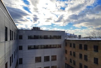 The image depicts a view of a building with a flat roof, seen from a vantage point that might be from another building across a courtyard. The sky is partially cloudy, and the sun is shining brightly, casting shadows onto the building's facade.
