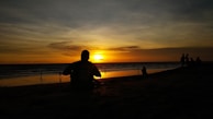 A peaceful sunset over the ocean with a silhouette of a girl sitting on the sand.