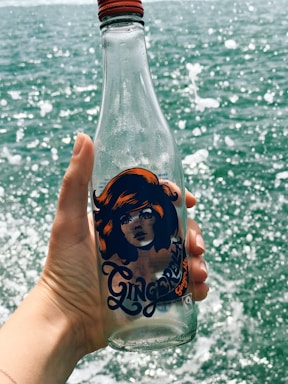 A hand holds a clear glass bottle with a colorful design featuring a woman's face and vibrant orange hair. The bottle has 'Ginger Brew' written on it. In the background, there is a body of water with waves splashing, creating a dynamic contrast.